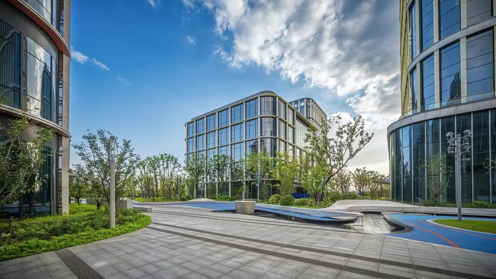 Modern glass facade commercial office buildings with landscaped pedestrian walkways and blue sky backdrop.